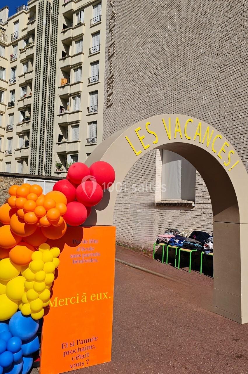 Arche beige avec l'inscription ’Les vacances’, décorée de ballons colorés, devant un immeuble résidentiel.