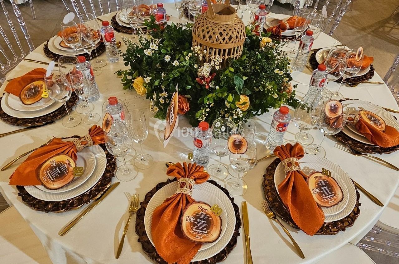 Table ronde élégamment dressée avec nappes blanches, serviettes orange, couverts dorés et centre de table floral.