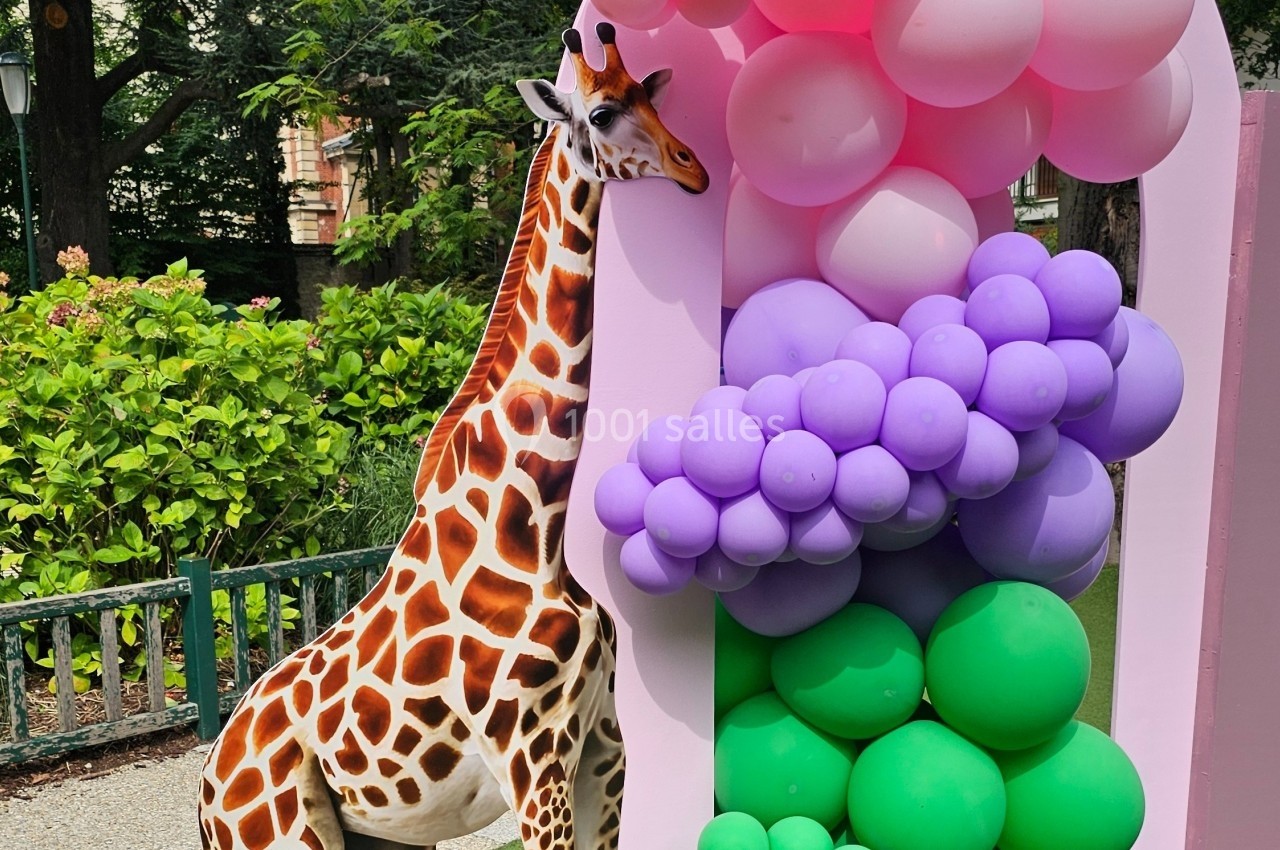 Une girafe en plastique à côté d'une structure décorative composée de ballons roses, violets et verts dans un parc.