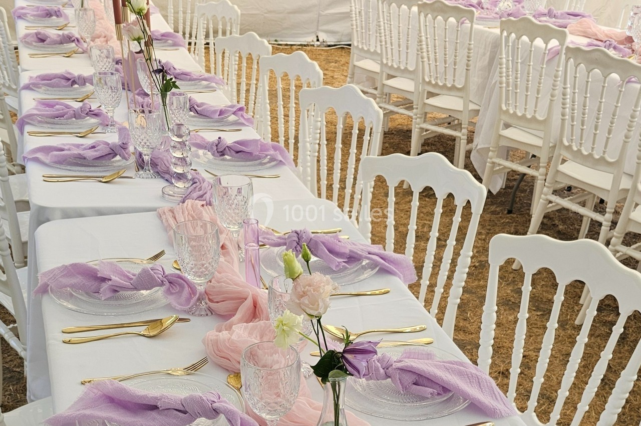 Tables dressées avec nappes blanches, décorations roses et violettes, couverts dorés et chaises blanches alignées.