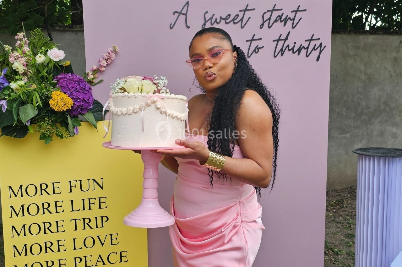 Une femme en robe rose tient un gâteau décoré de perles devant un décor floral et des panneaux colorés.
