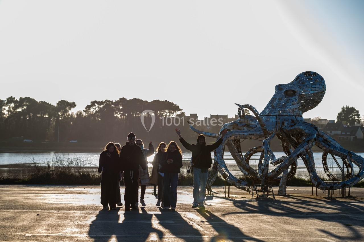 Un groupe de personnes marche près d'une sculpture métallique en forme de pieuvre, avec un paysage naturel en arrière-plan.