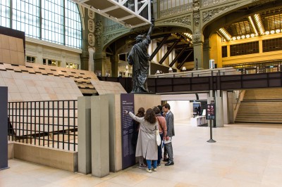 Des visiteurs lisent des informations près d'une réplique de la statue de la Liberté dans un espace intérieur lumineux.