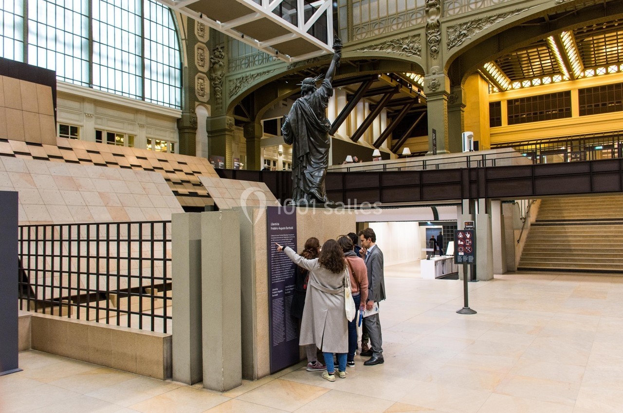 Des visiteurs lisent des informations près d'une réplique de la statue de la Liberté dans un espace intérieur lumineux.