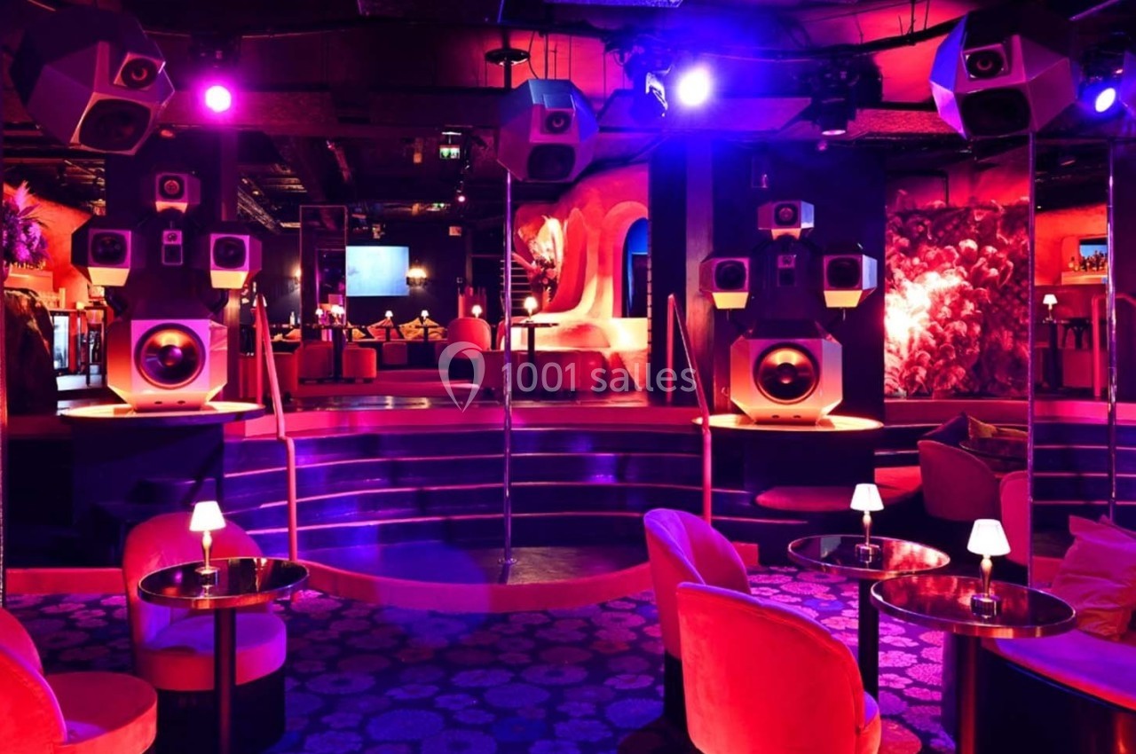 Salle de club colorée avec éclairage violet, sièges rembourrés, tables basses et haut-parleurs suspendus.
