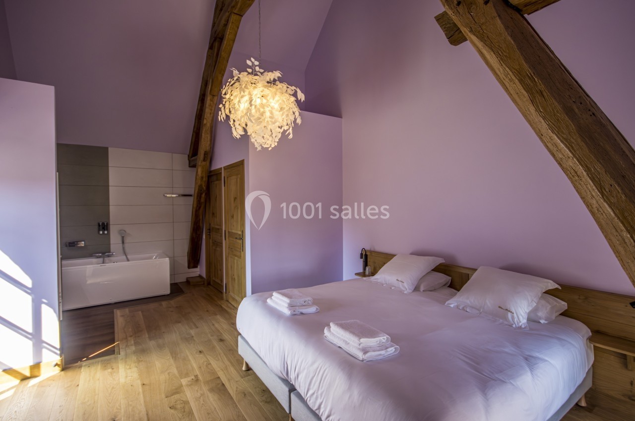 Chambre avec poutres apparentes, lit double, baignoire moderne et murs peints en violet clair.