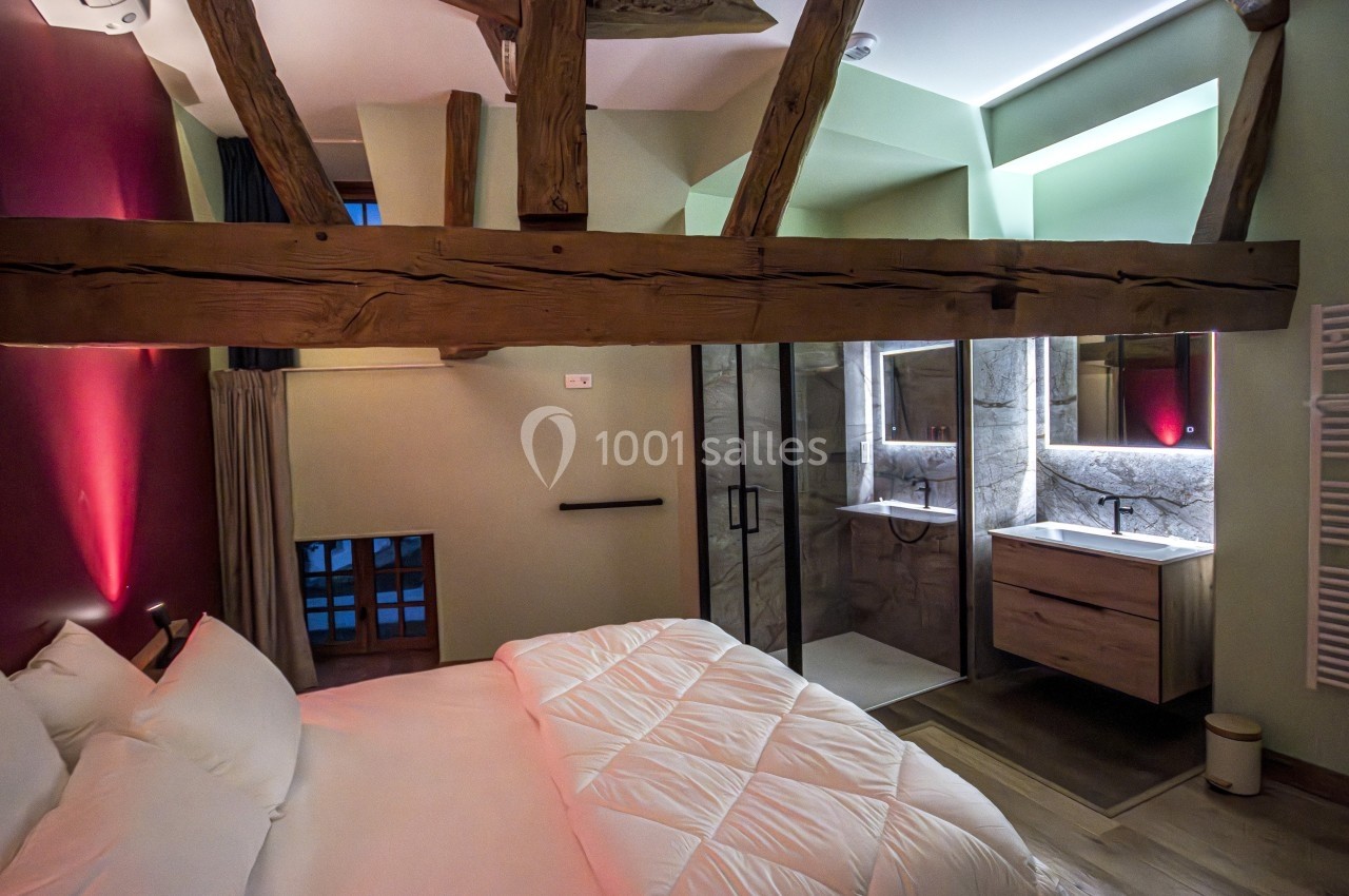 Chambre avec poutres apparentes, lit double, éclairage tamisé et salle de bain attenante avec douche moderne.