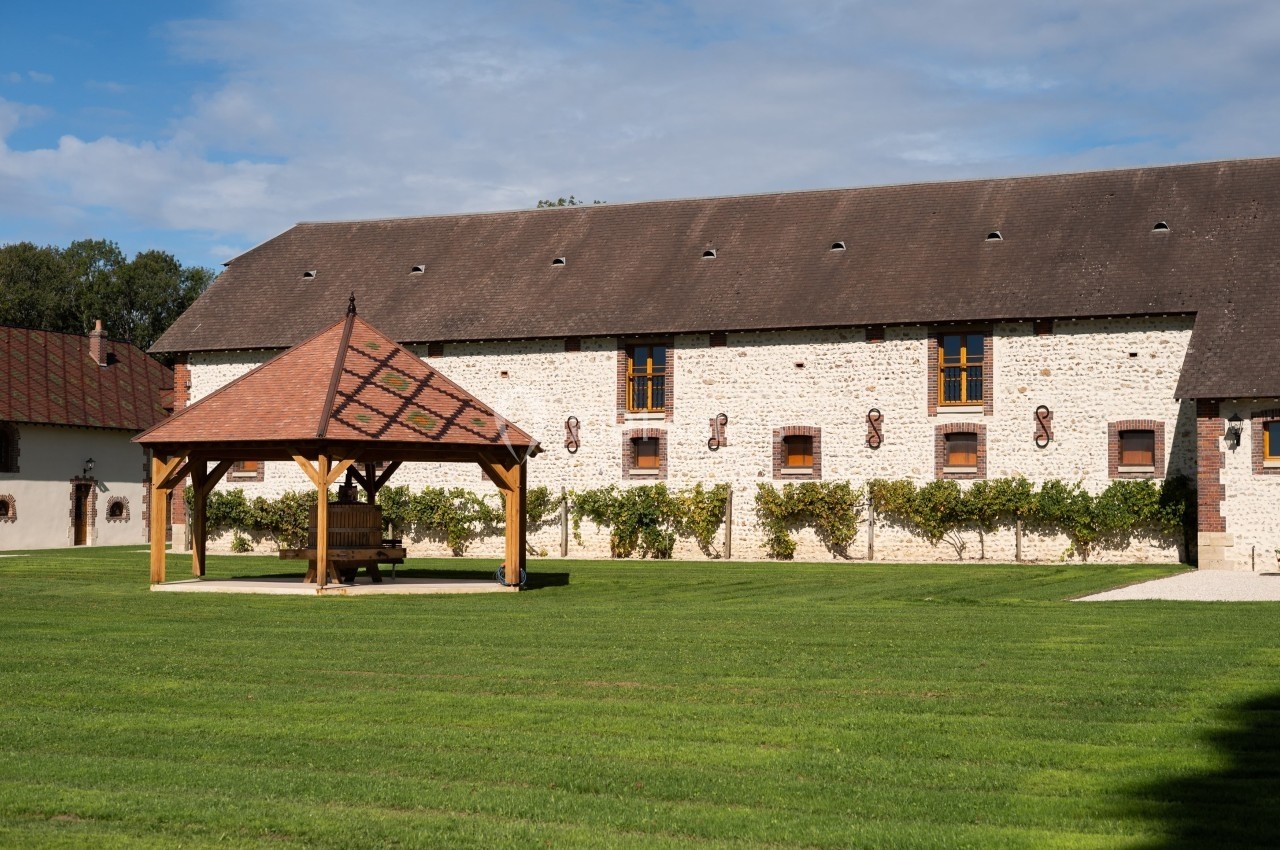 Bâtiment en pierre avec toit en ardoise, pergola en bois et pelouse bien entretenue sous un ciel partiellement nuageux.