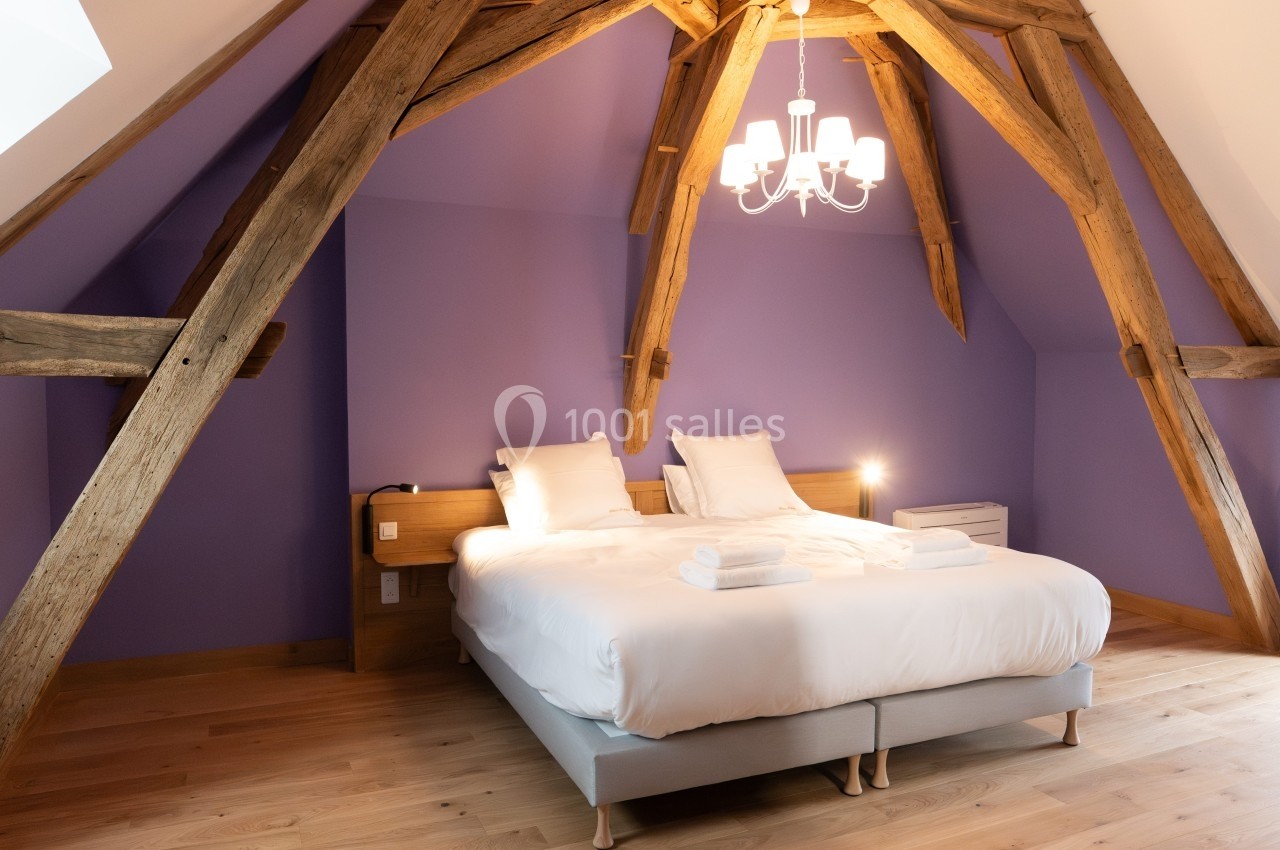 Chambre mansardée avec poutres apparentes, lit double, linge de lit blanc et mur violet, éclairée par un lustre.