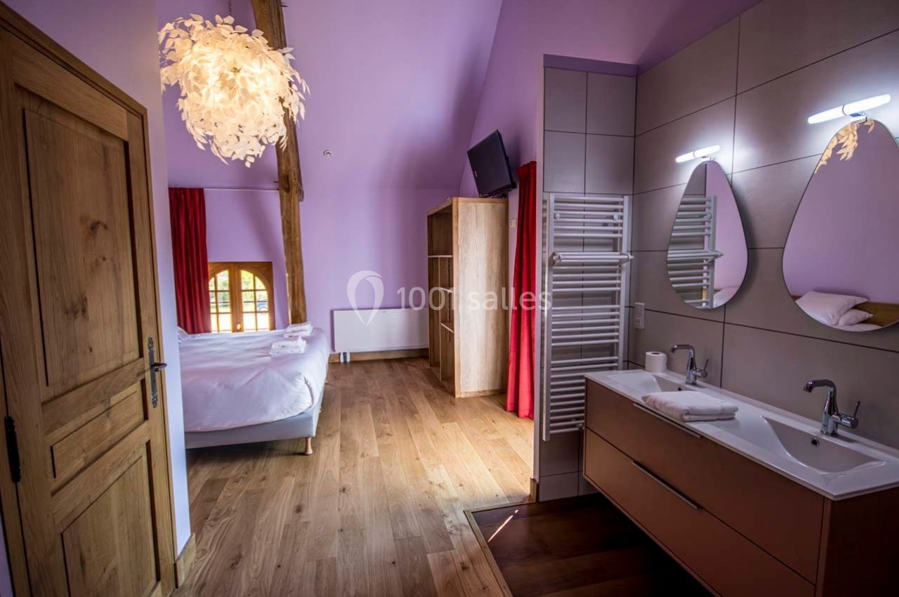 Chambre lumineuse avec lit double, parquet, salle de bain attenante équipée de deux lavabos et murs peints en violet.