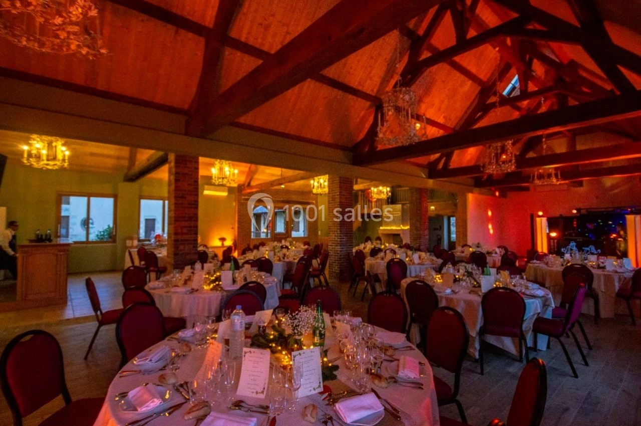 Salle de réception chaleureuse avec tables rondes dressées, éclairage tamisé et poutres apparentes en bois.