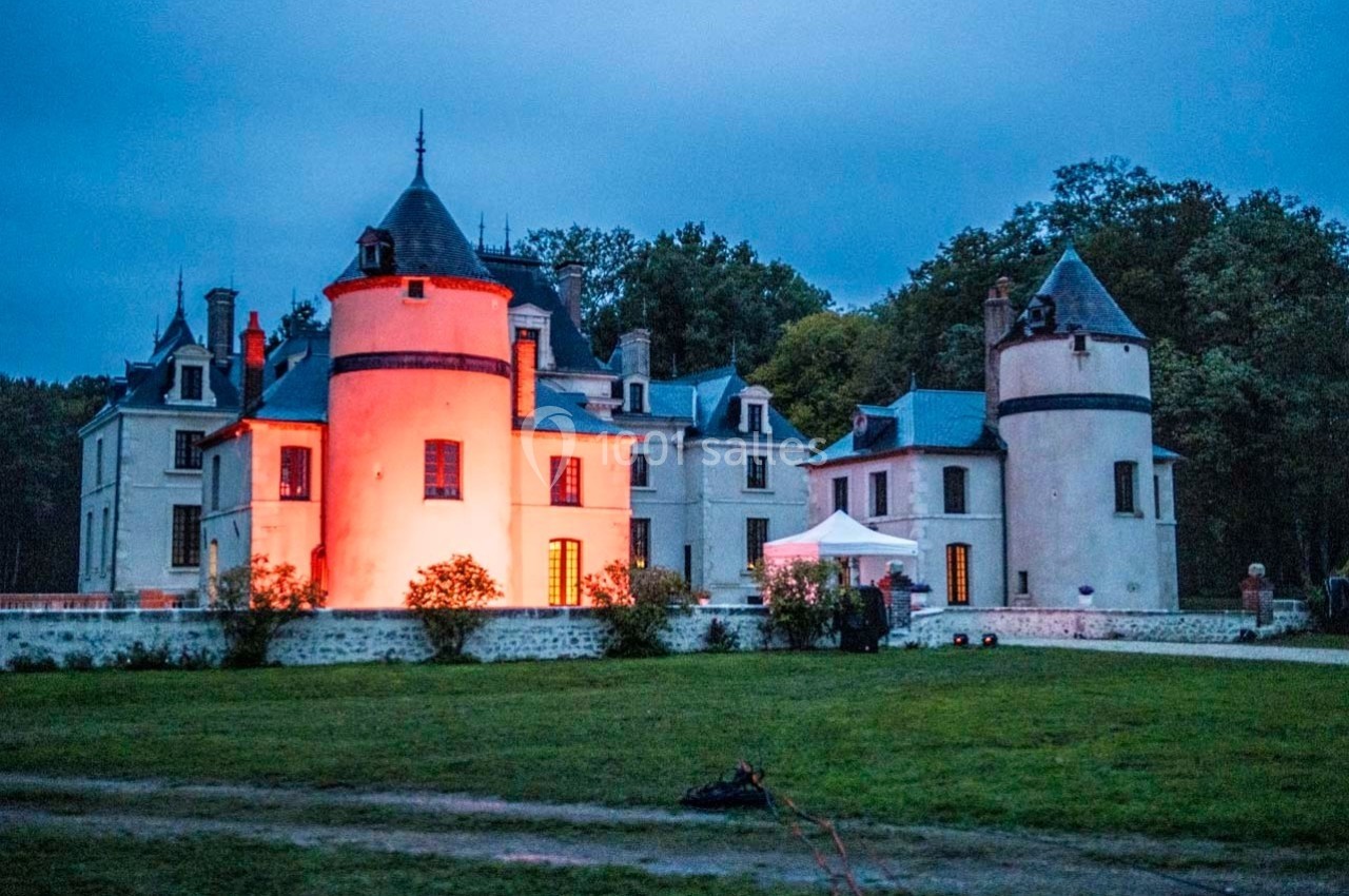 Château éclairé par des lumières rouges au crépuscule, entouré de verdure et d'arbres en arrière-plan.
