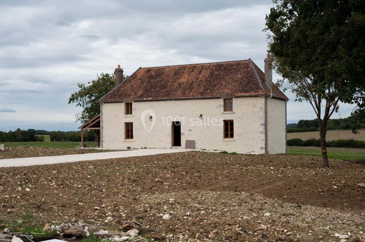 Maison en pierre blanche avec toit en tuiles rouges, entourée de terrain dégagé et d'arbres sous un ciel nuageux.