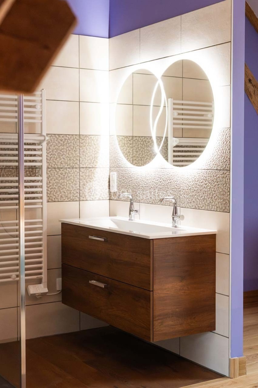 Meuble de salle de bain en bois avec double vasque, miroir rond éclairé et carrelage mural texturé.