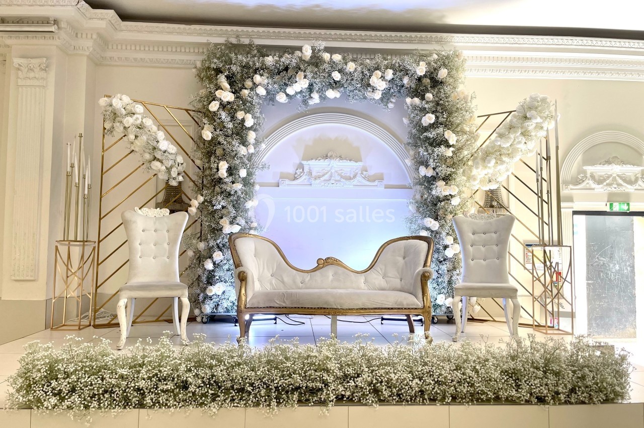 Décor de cérémonie avec canapé blanc, deux fauteuils assortis, fleurs blanches et arches dorées sur une estrade.