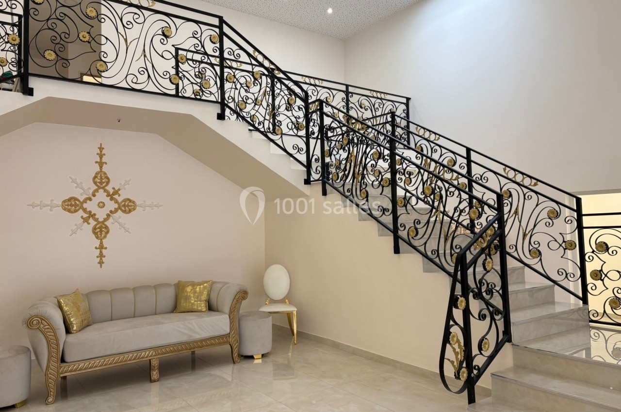 Escalier en métal noir orné de motifs dorés, avec un canapé beige et des coussins dorés dans un intérieur lumineux.