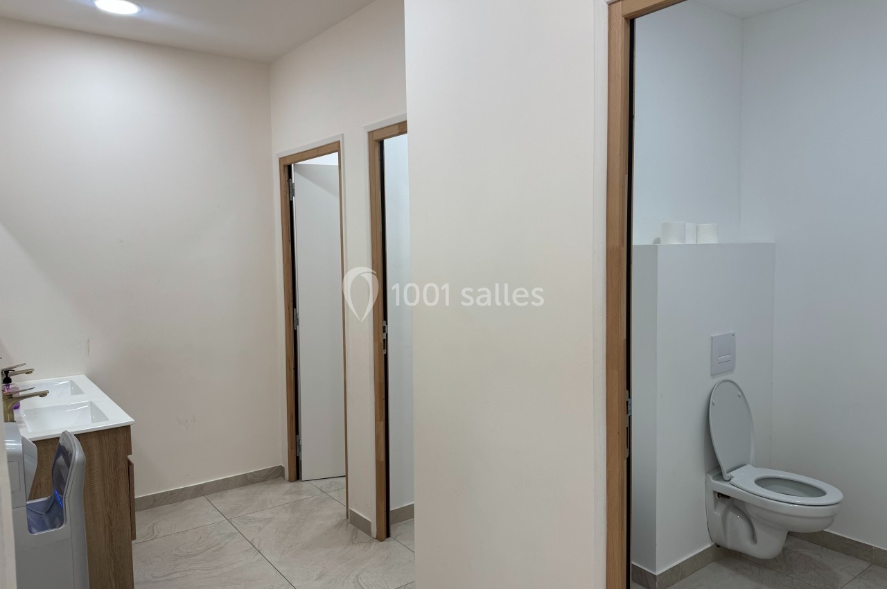 Vue d'un espace sanitaire avec un lavabo à gauche et des toilettes séparées par des portes à droite.