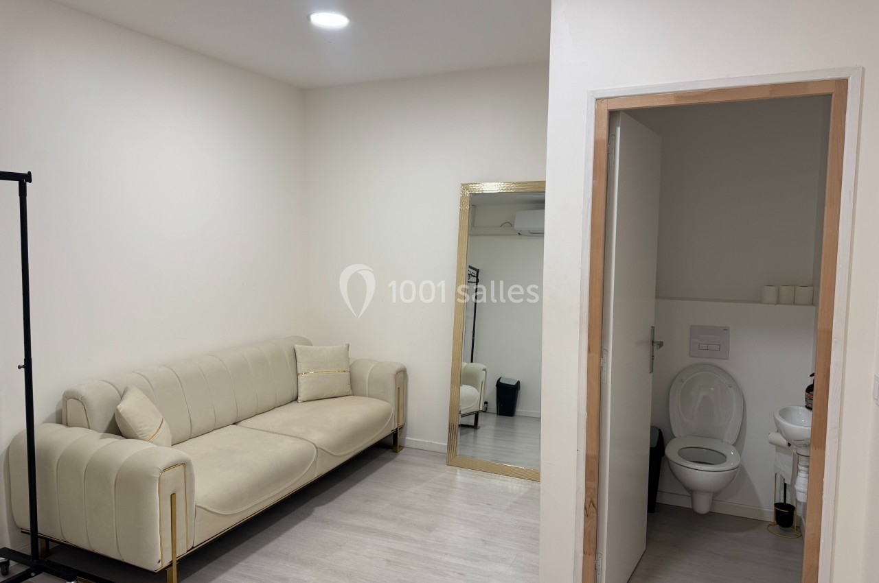 Salle lumineuse avec canapé beige, miroir doré, portant vide et porte ouverte donnant sur des toilettes.
