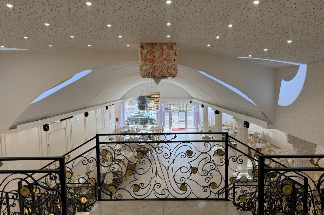 Salle de réception lumineuse avec plafond voûté, lustre central, tables dressées et balustrade décorative au premier plan.