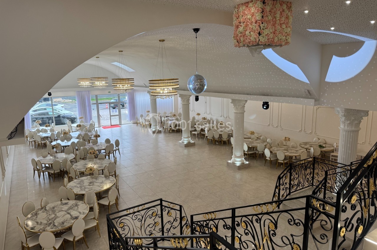 Salle de réception spacieuse avec tables rondes, chaises élégantes, lustres suspendus et escalier décoratif en fer forgé.
