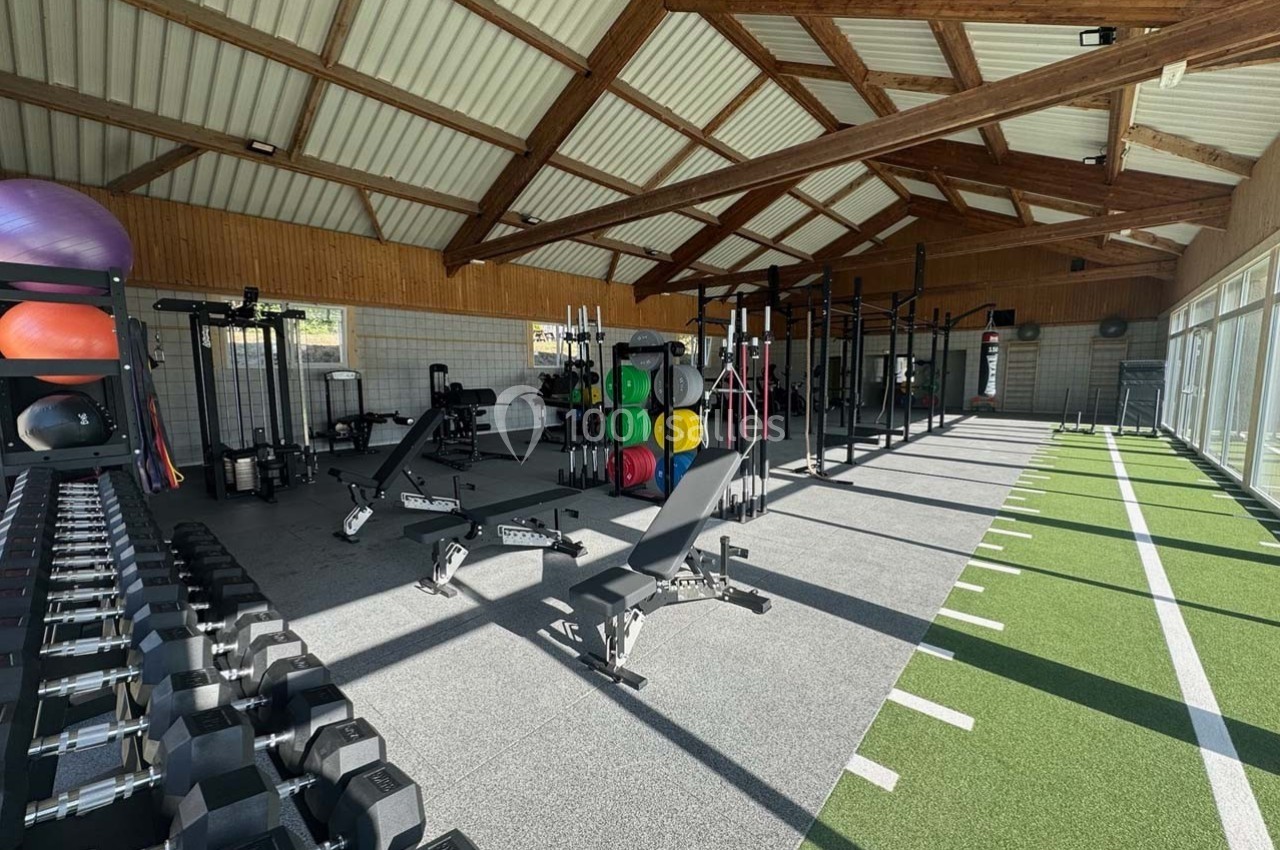 Salle de sport lumineuse avec équipements de musculation, haltères, bancs et espace de gazon synthétique.
