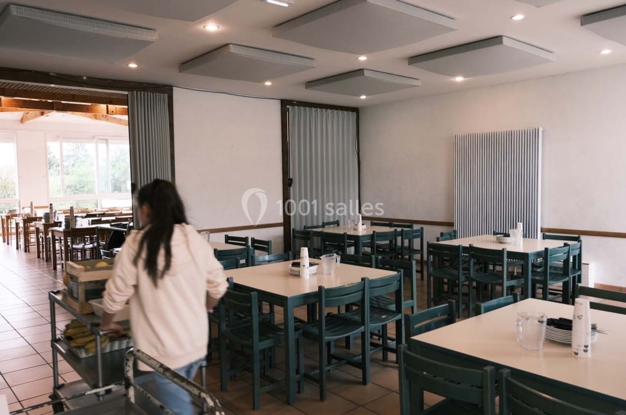 Une personne de dos range un chariot dans une salle de restaurant lumineuse avec des tables et des chaises alignées.