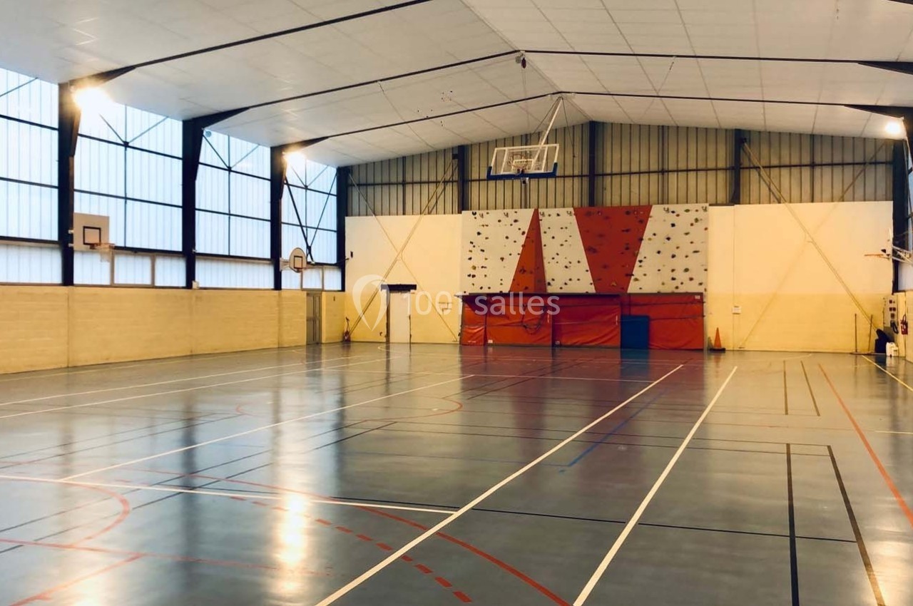 Salle de sport avec terrain de basketball, murs d'escalade et éclairage naturel provenant de grandes fenêtres latérales.