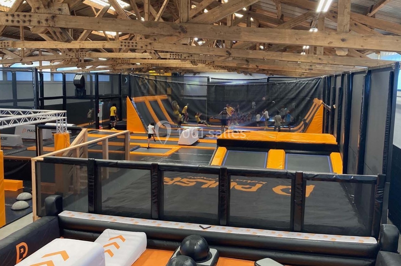 Salle intérieure avec trampolines, structures gonflables et participants en activité sous une charpente en bois.