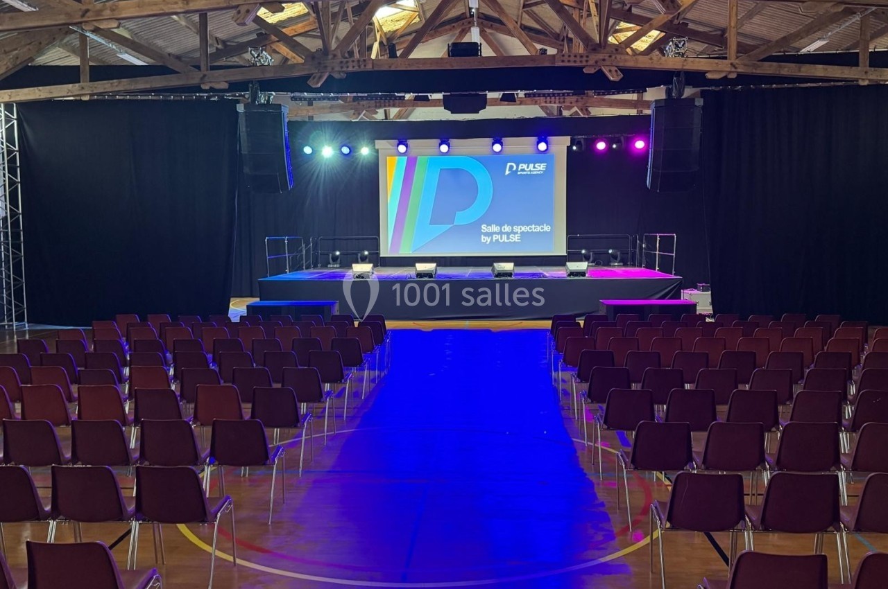 Salle de spectacle avec scène éclairée et rangées de chaises vides dans un espace à charpente apparente.