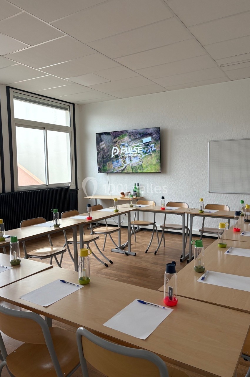 Salle de réunion lumineuse avec tables disposées en U, chaises, fournitures de bureau et écran affichant ’Pulse’.
