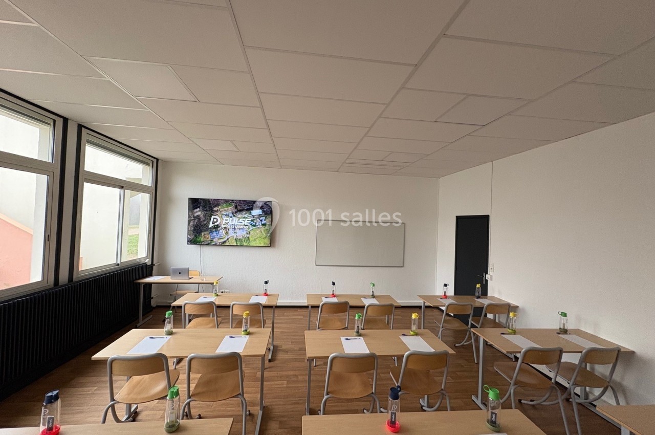 Salle de classe lumineuse avec des tables en bois, des chaises, un tableau blanc et un écran mural allumé.