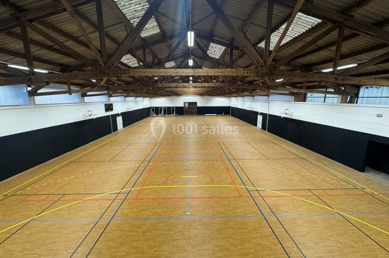 Salle de sport intérieure avec parquet, marquages au sol pour différents sports et structure en bois apparente.
