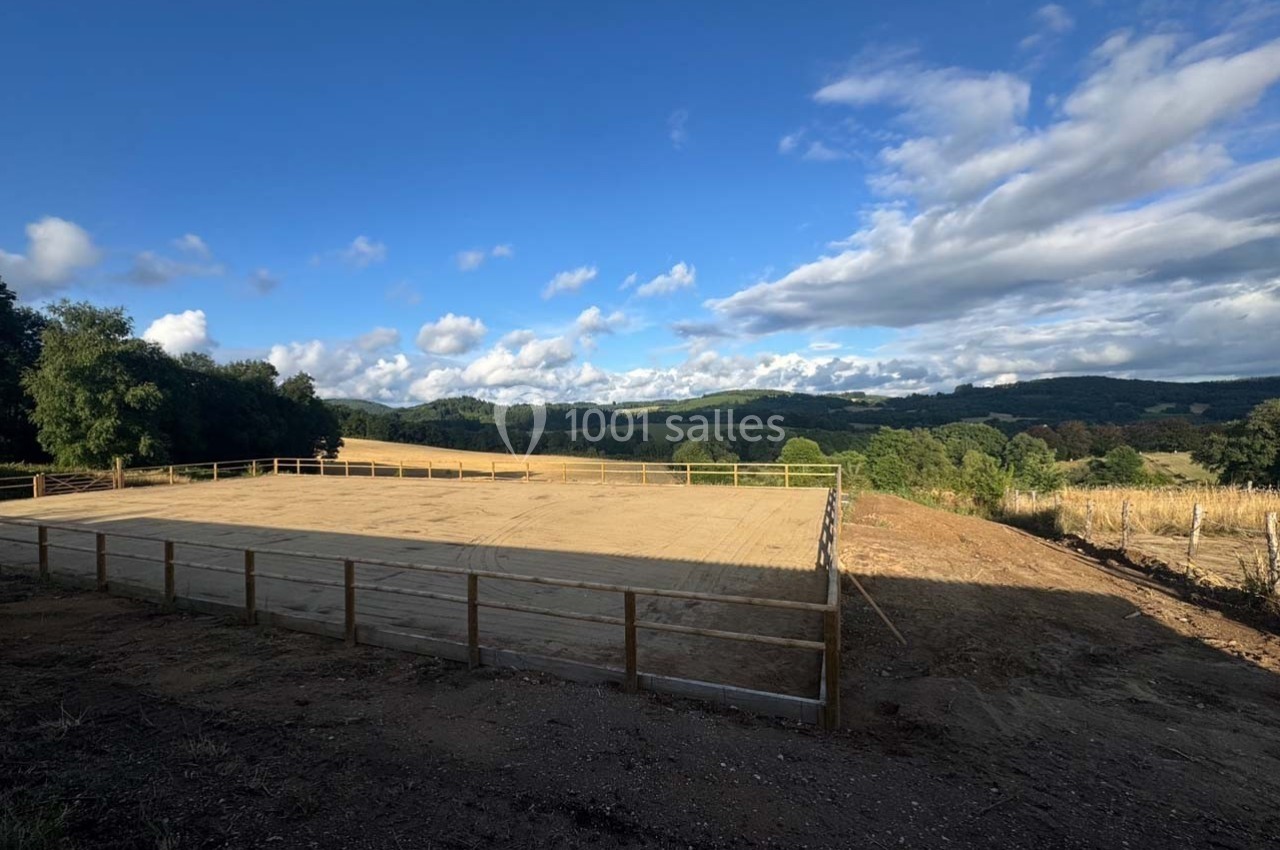 Vue d'une carrière équestre en sable entourée de barrières, avec un paysage vallonné et un ciel partiellement nuageux.