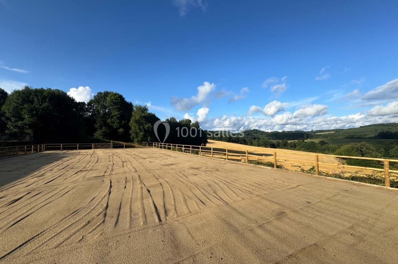 Vue d'une carrière équestre en sable entourée de clôtures en bois, avec des champs et des arbres à l'horizon.