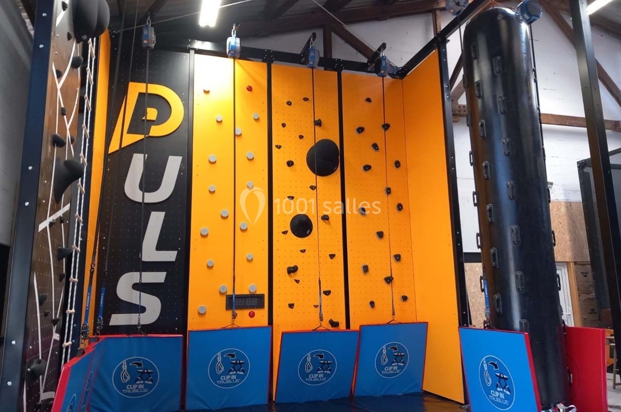 Mur d'escalade intérieur avec prises variées, panneaux jaunes et noirs, et tapis de protection au sol.