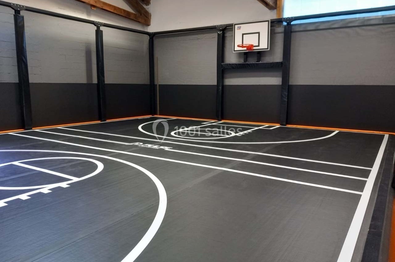 Terrain de basketball intérieur avec sol noir, lignes blanches et panneau de basket fixé sur un mur.