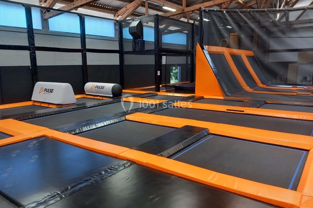Salle intérieure avec trampolines orange et noirs, structures inclinées et filets de protection sous un toit en bois.