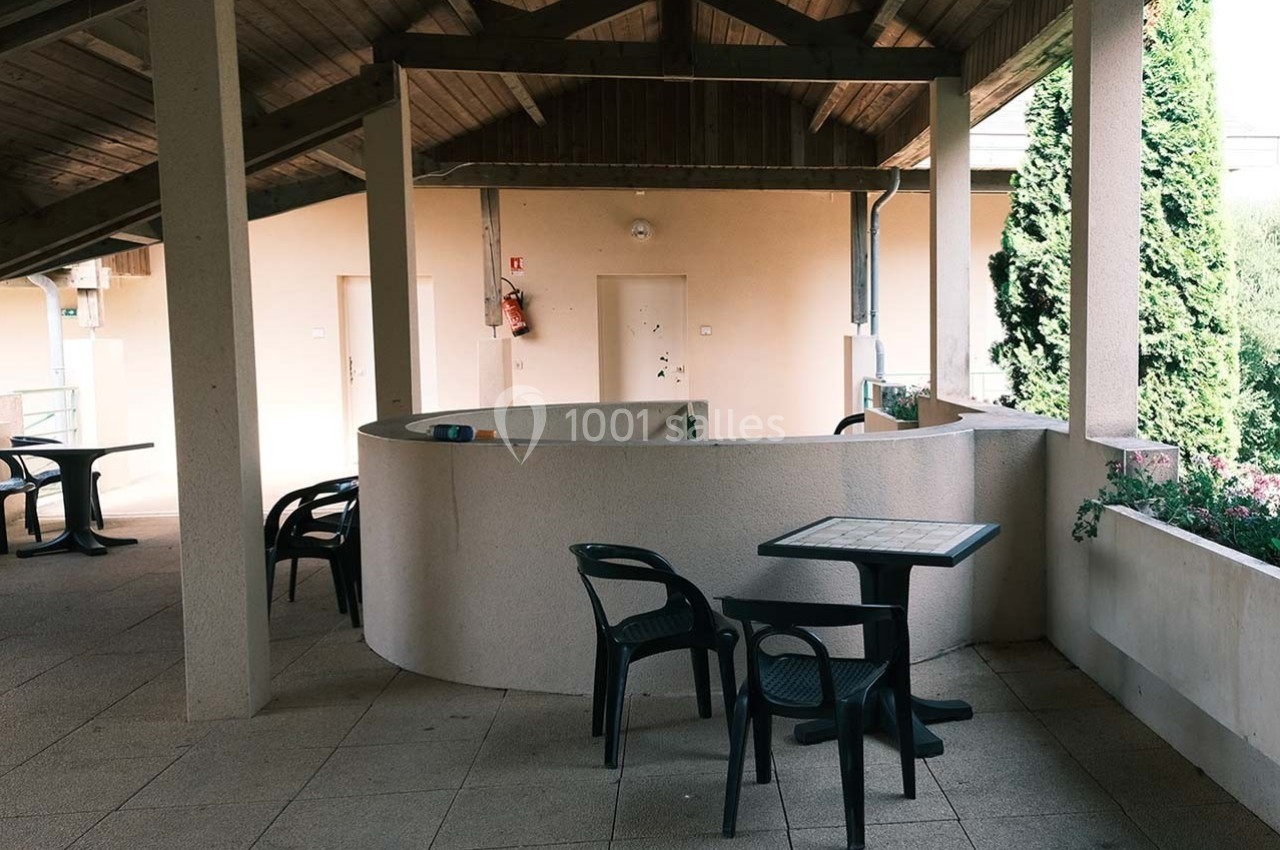 Terrasse couverte avec tables et chaises en plastique noir, entourée de murs clairs et végétation en arrière-plan.