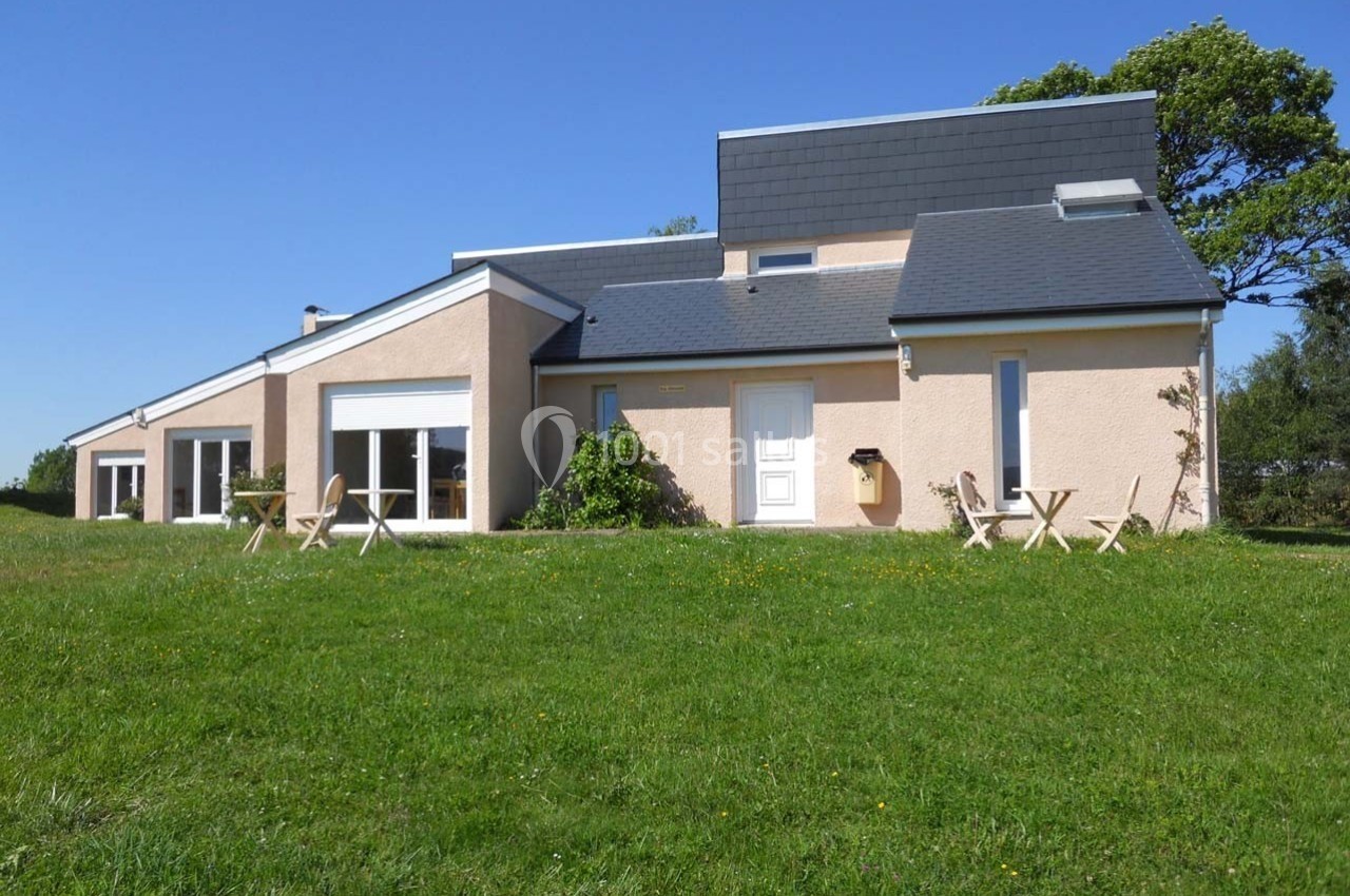 Maison moderne beige avec toit en ardoise, grandes baies vitrées et jardin verdoyant sous un ciel bleu.
