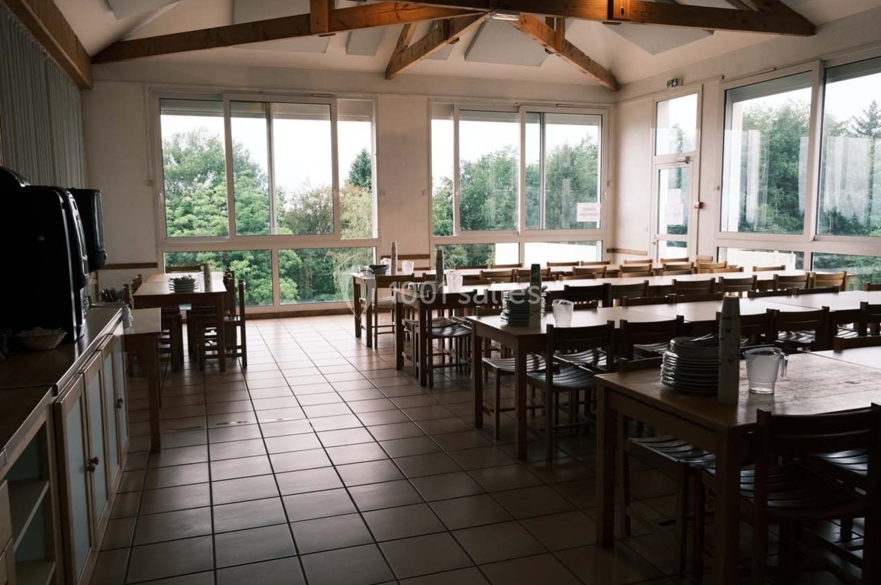 Salle à manger lumineuse avec de grandes fenêtres, des tables en bois alignées et de la vaisselle empilée.