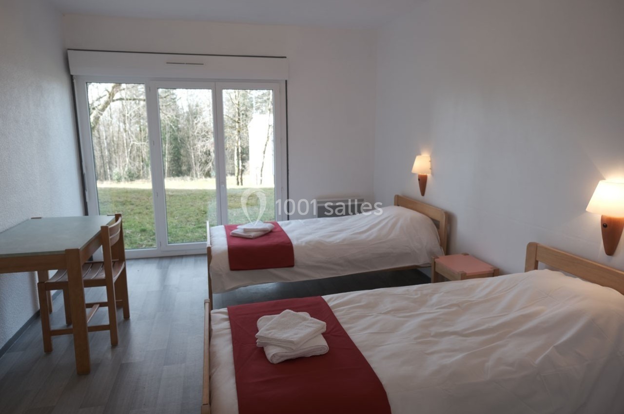 Chambre lumineuse avec deux lits simples, table en bois, et grande baie vitrée donnant sur un espace vert.