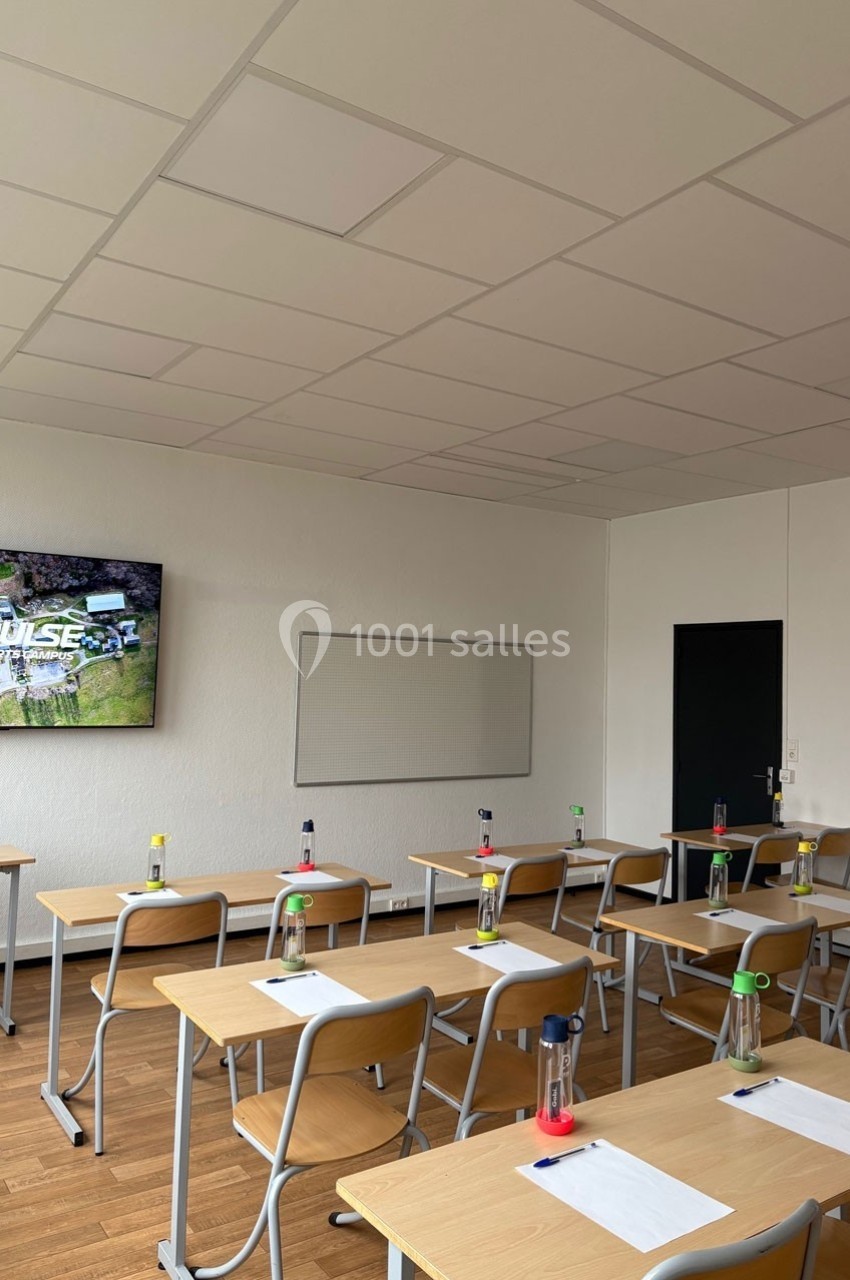 Salle de classe lumineuse avec des bureaux alignés, des bouteilles d'eau et un écran allumé au mur.