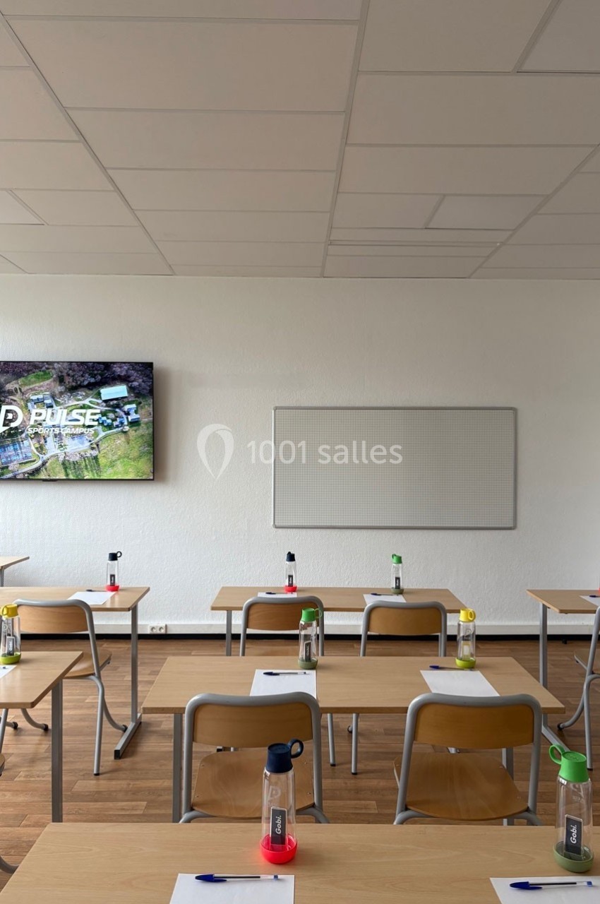 Salle de classe lumineuse avec des bureaux alignés, des bouteilles colorées, un tableau blanc et un écran allumé au mur.