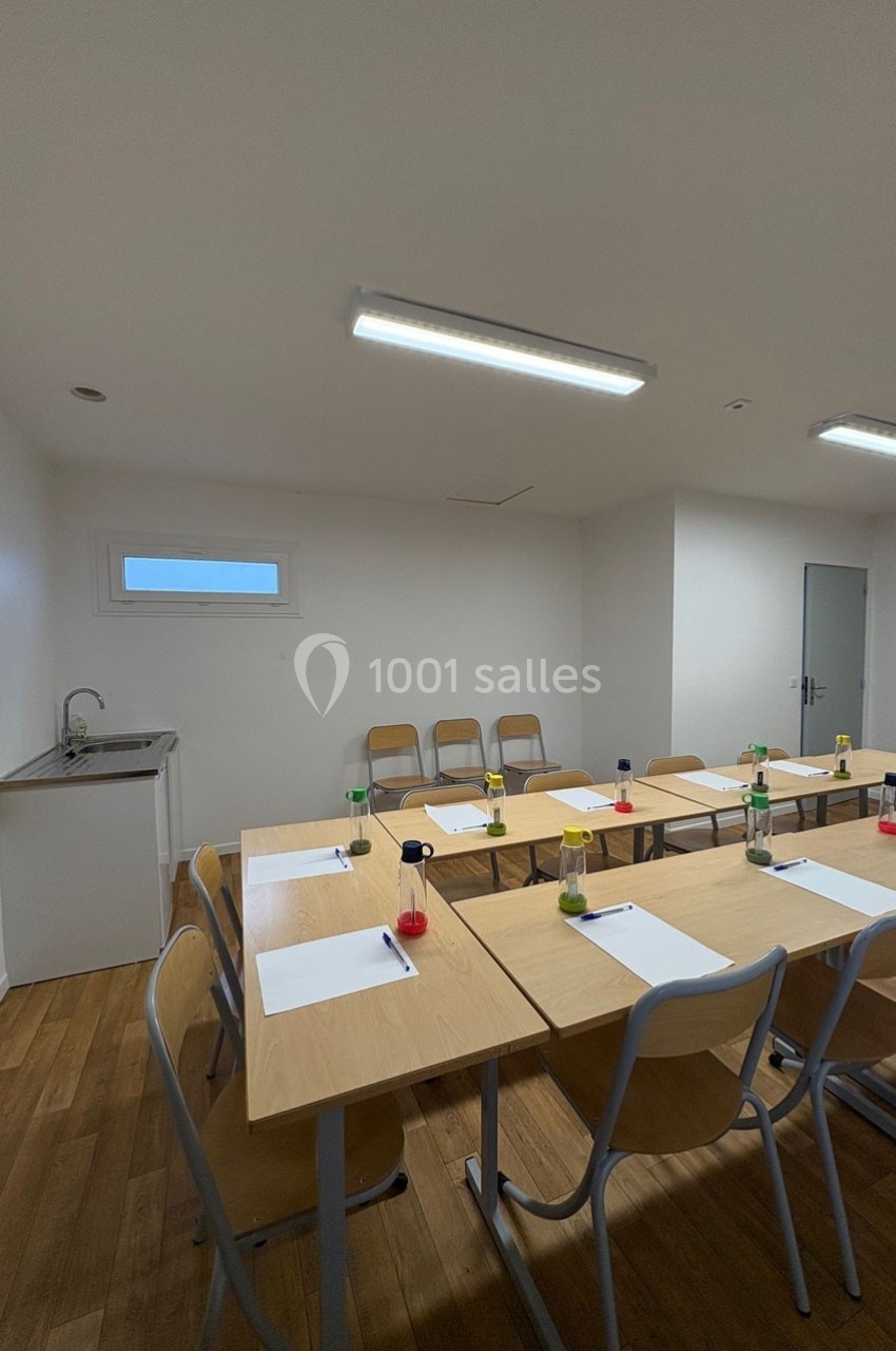 Salle de réunion lumineuse avec tables en bois disposées en U, chaises, feuilles, stylos et un coin évier.