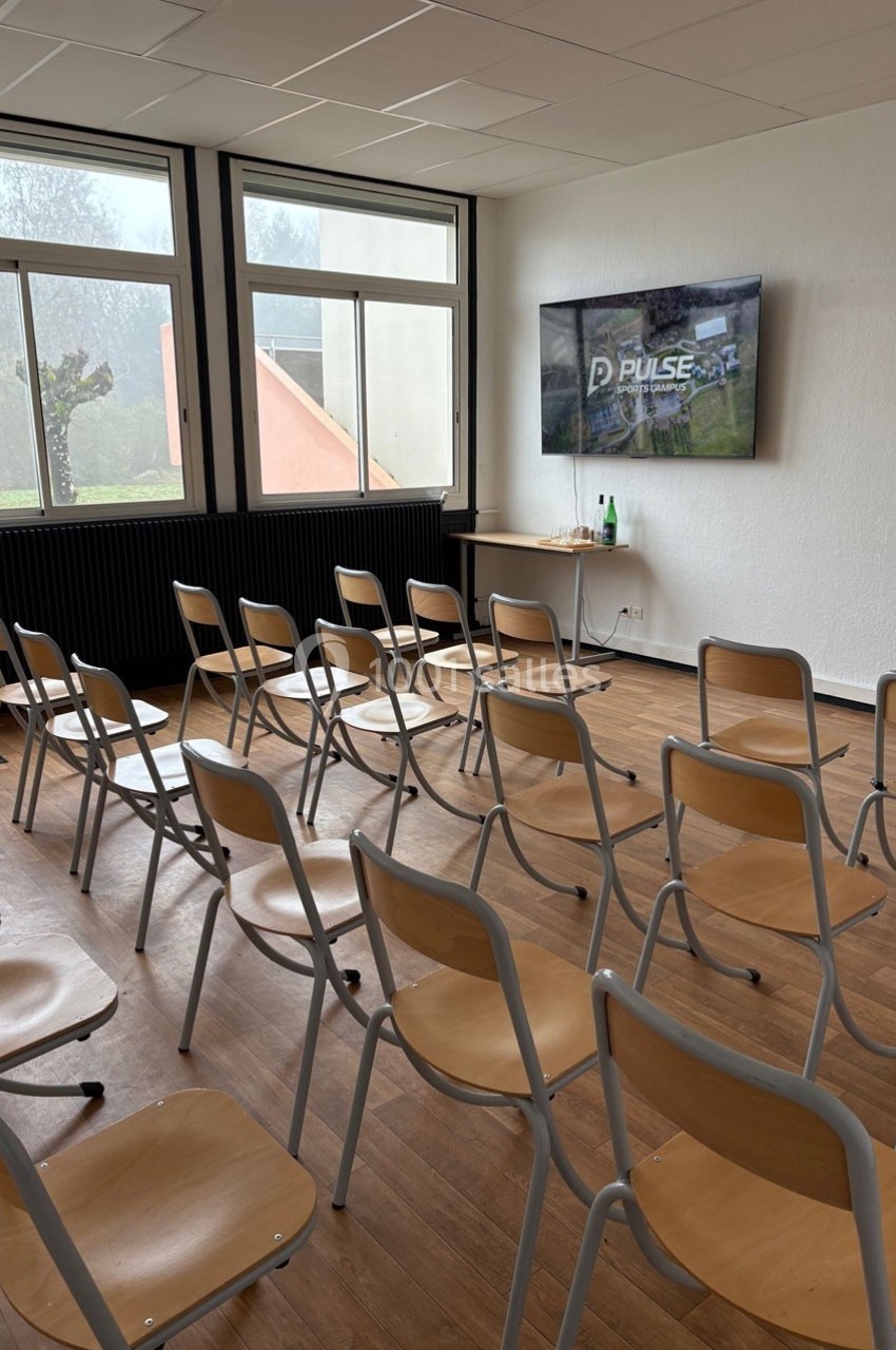Salle de réunion lumineuse avec des rangées de chaises en bois, un écran mural allumé et une vue sur l'extérieur.