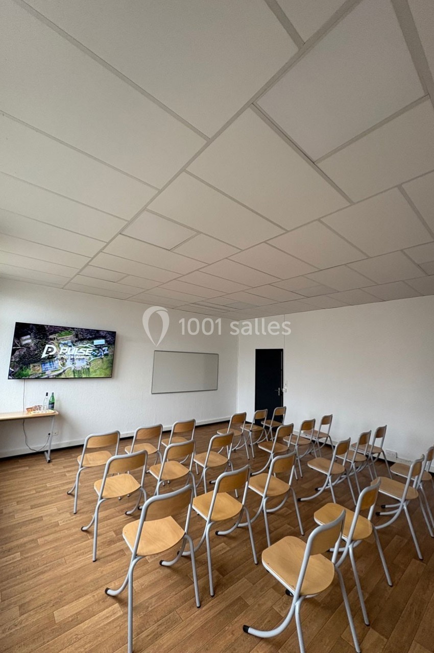 Salle de réunion avec chaises alignées, écran mural allumé, tableau blanc et sol en parquet.