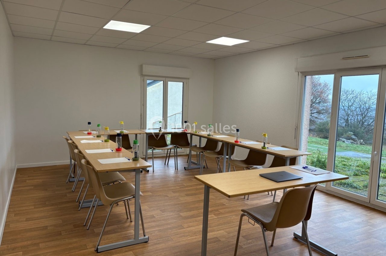 Salle de réunion lumineuse avec tables en U, chaises, bouteilles d'eau et vue sur un extérieur arboré.
