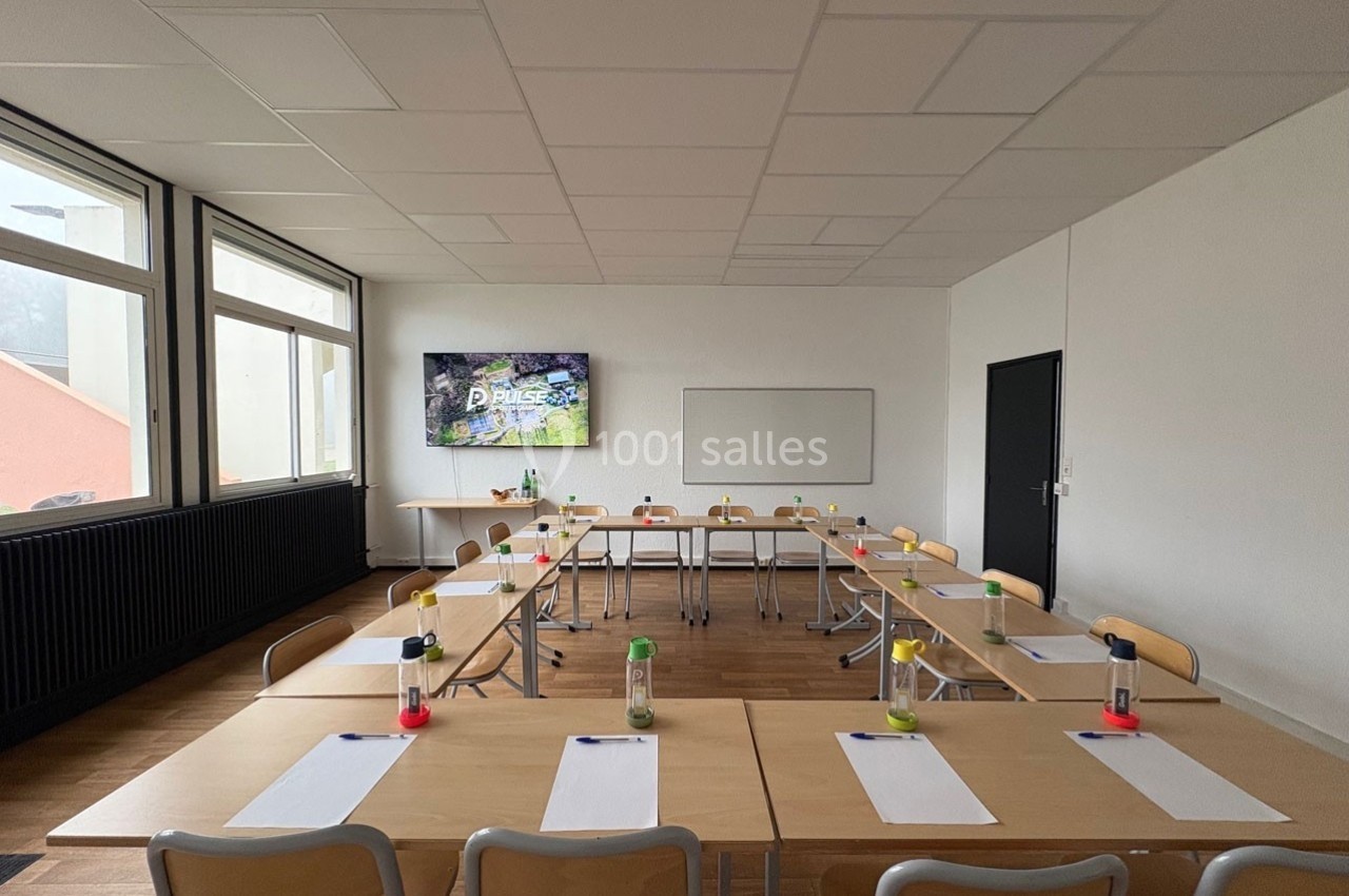 Salle de formation lumineuse avec tables disposées en U, écran, tableau blanc et fournitures sur les bureaux.