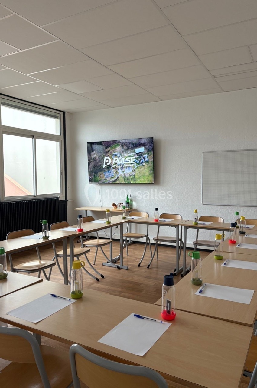 Salle de réunion lumineuse avec des tables disposées en U, équipée d'un écran mural et de fournitures de bureau.