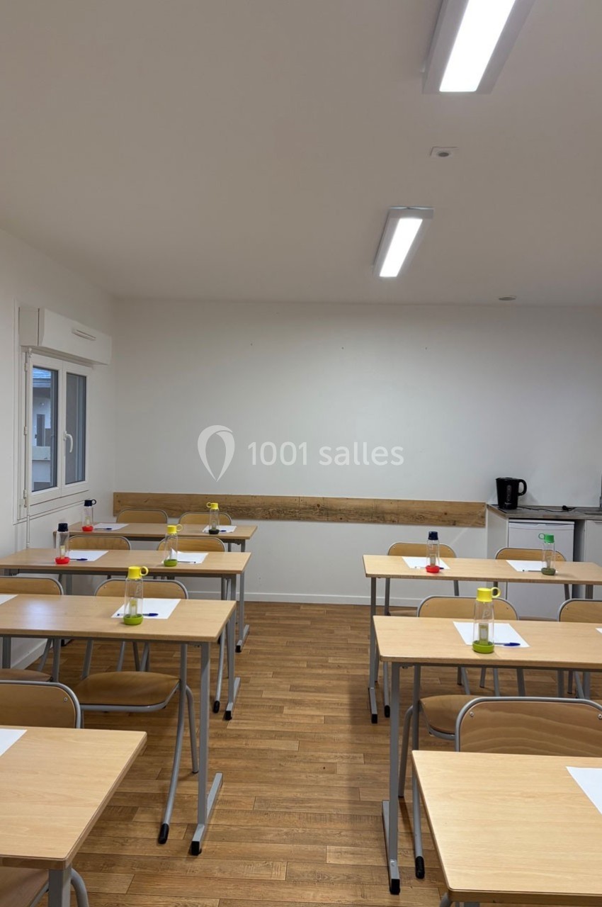 Salle de classe vide avec des bureaux en bois, des feuilles et des stylos, éclairée par des néons au plafond.